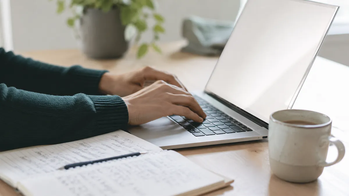 Mains sur le clavier d'un portable, à côté d'un carnet annoté et d'une tasse de café